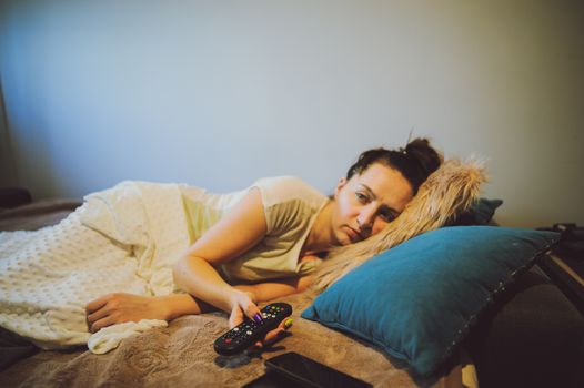 sick woman lying in bed with headache holding tv remote in hand