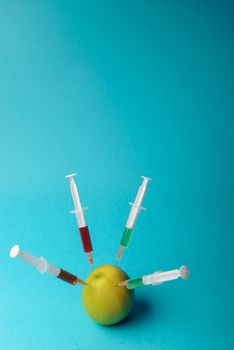 Chemical additives in food or genetically modified fruit concept. Green apple with syringes of chemicals. Isolated on blue background.