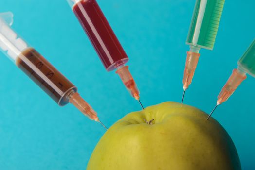 Chemical additives in food or genetically modified fruit concept. Green apple with syringes of chemicals. Isolated on blue background.
