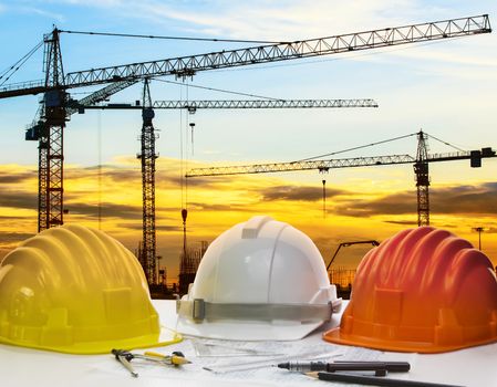 safety helmet and engineering plan with drawing instrument on engineer working table against crane construction and beautiful sun set scene use for construction and land development topic