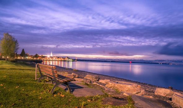 Colorful view over the Bodensee lake as the sun starts rising, with a wooden bench on its shore. Image captured in Friedrichshafen, Germany.