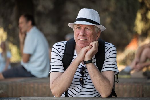 A male senior tourist traveling alone. Wearing a white summer hat, backpack and a white t-shirt with black stripes.  He is pensive and contented