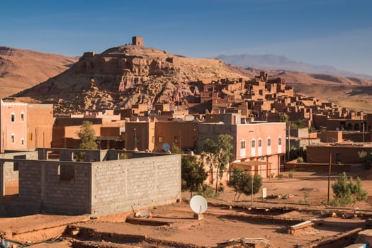 View on a hill with an  old medieval city, built in red color. New building in the foreground, Atlas mountains in the background. Blue morning sky.