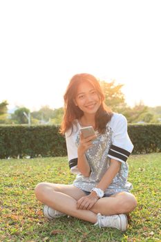 young teen woman sitting on grass field with smart phone in hand and toothy smiling with happy emotion 