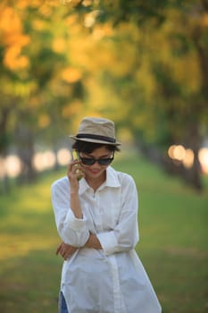 beautiful young woman wearing sun glassea nd straw hat talking on mobile phone with happiness emotion against blurry yellow flower in park use for people and modern life in digital connecting technology