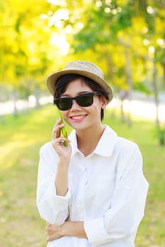 young beautiful woman wearing sun glasses and fashion straw hat talking on mobile phone with happy face toothy smiling against blur of yellow flowers blooming in park