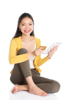 Full length portrait of casual Asian woman sitting on floor using touch screen tablet pc for online shopping and making payment, isolated on white background.