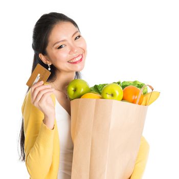 Happy young Asian female shopper, hands holding shopping bags filled with groceries and showing credit card, isolated standing on white background.