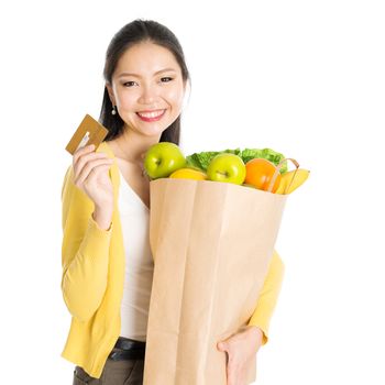 Happy young Asian female shopper, hands holding shopping bags full of groceries and showing credit card, isolated standing on white background.