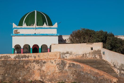 Mosque built on a hill on the coast of Atlantic Ocean in a moroccan city Sidi Ifni. Colorful details of the mosque. Bright blue sky. Old stairs and fence.