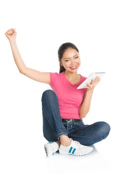 Full body young Asian girl in pink shirt using digital tablet computer and cheering, seated on floor, full length isolated on white background.
