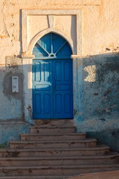 Entrance to the house from a precipitous street. Several stairs to the blue ornate gate, enlightened by the late afternoon sunlight. Sidi Ifni, Morocco.