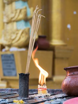Joss sticks and a bundle of candles burning at dawn in front of a golden stupa at the Shwedagon Pagoda in Yangon, Myanmar. Shallow depth of field with the joss sticks in focus.