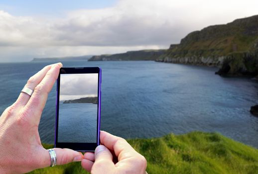 View over the mobile phone display during taking a picture of coast and ocean in Northern Ireland. Holding the mobile phone in hands and taking a photo. Focused on mobile phone screen. 