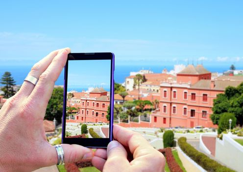 View over the mobile phone display during taking a picture of castle and garden Victoria in La Orotava, Tenerife. Holding the mobile phone in hands and taking a photo. Focused on mobile phone screen. 