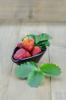 fresh strawberry fruits in black bowl with green leaves over wooden background