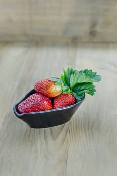 fresh strawberry fruits in black bowl with green leaves over wooden background