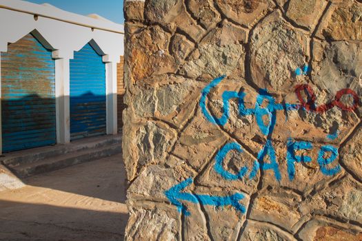 Stone wall with a hand written "cafe" in arabian and latin script and an arrow. In the background closed shops. Sidi Ifni beach, Morocco.