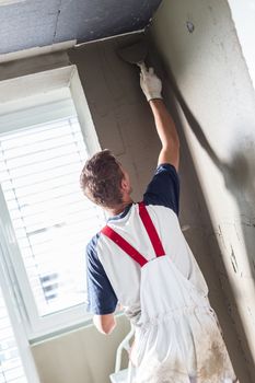 Thirty years old manual worker with wall plastering tools renovating house. Plasterer renovating indoor walls and ceilings with float and plaster.