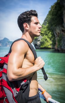 Portrait of young handsome bare chested brunet man looking away against seascape on a boat or ship, with rucksack on shoulder
