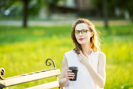 Portrait of lovely urban girl with paper cup in her hands. Happy smiling woman walking in a city park. Fashionable blonde girl wearing spectacles