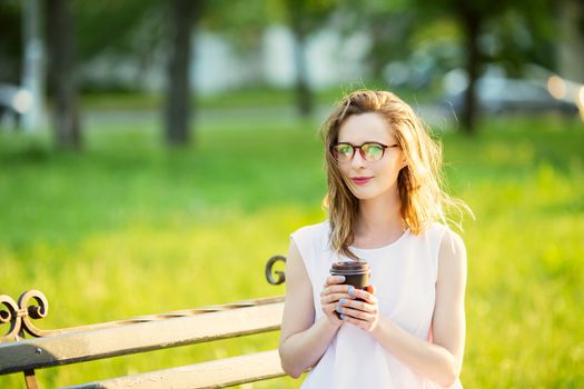 Portrait of lovely urban girl with paper cup in her hands. Happy smiling woman walking in a city park. Fashionable blonde girl wearing spectacles