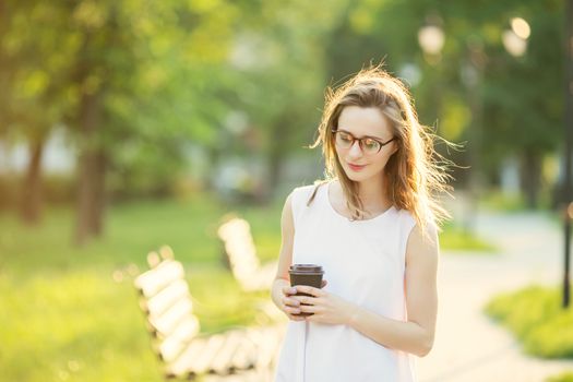 Portrait of lovely urban girl with paper cup in her hands. Happy smiling woman walking in a city park. Fashionable blonde girl wearing spectacles