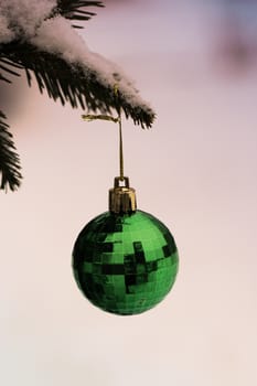 Green Christmas ball hanging on natural branch of pine in snowy weather.Christmas toy in the shape of a ball hanging on a spruce branch