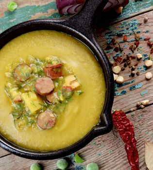 Pea Soup with Smoked Sausages in Black Iron Cast with Yellow and Green Pea, Potato, Greens and Spices closeup on Wooden background. Top View