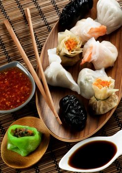 Assorted Dim Sum in Wooden Bowls, Red Chili and Soy Sauces and Chopsticks closeup on Straw Mat background