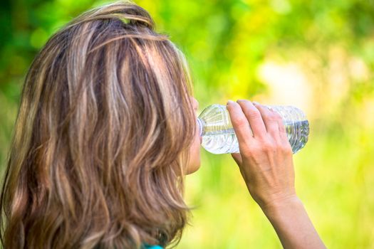 A blonde lady drinking from a bottled water outside on a sunny afternoon