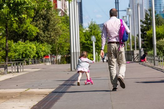 A father walking and young happy daughter skatingalong the street in the afternoon.