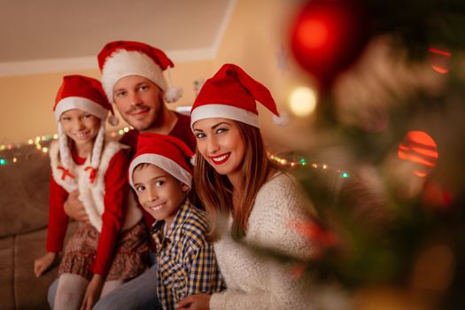 Beautiful smiling family are gathered together by a Christmas tree at the home. Looking at camera and wearing Santa hats. 