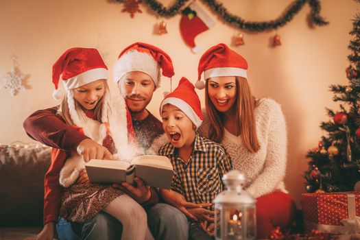 Beautiful smiling family of four with santa hat reading book at the home.