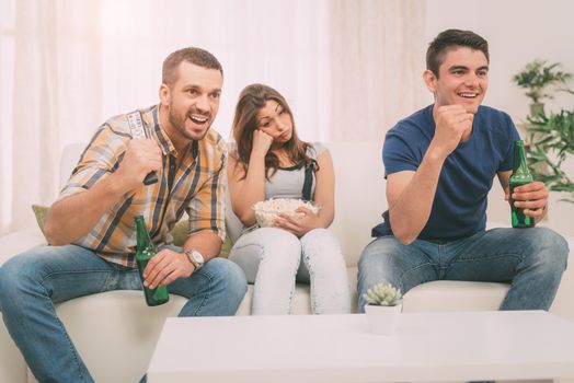 Three friends hanging out in an apartment and watching a football game together. Two man cheering and enjoying, girl is bored.