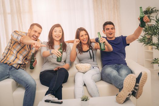 Four cheerful friends hanging out in an apartment and watching a football game together. They cheering, drinking beer and eating popcorn.