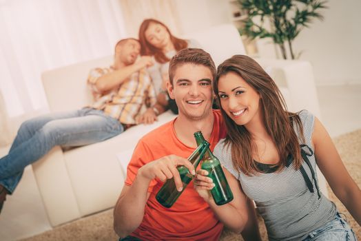 Young happiness couple enjoying at home party and toasting with beer. Their friends in the background. Selectiv focus. Focus on foreground. Looking at camera.