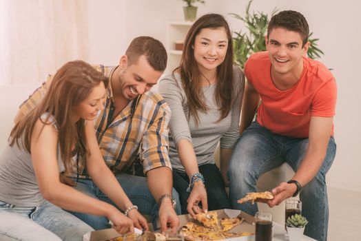 Four cheerful friends hanging out in an apartment and eating pizza.