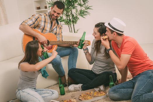 Four cheerful friends enjoing with guitar in an apartment. They drinking beer and eating pizza.