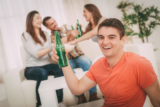 Close up of a young smiling man at home party with beer and cheers. His friends in the background. Selectiv focus. Focus on foreground.