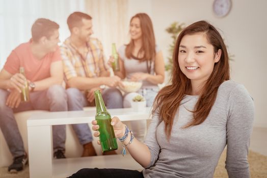 Close up of a young Japanese girl smiling at home party with beer and cheers. Her friends in the background. Selectiv focus. Focus on foreground.