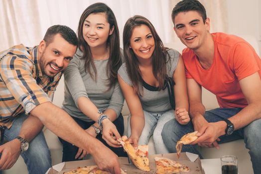 Four cheerful friends hanging out in an apartment. They eating pizza and looking at camera.