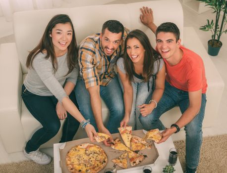 Four cheerful friends hanging out in an apartment. They eating pizza and looking at camera.