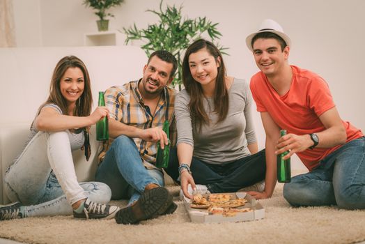 Four cheerful friends hanging out in an apartment. They drinking beer and eating pizza. Looking at camera.