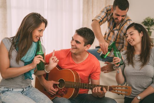 Four cheerful friends enjoing with guitar and beer in an apartment.