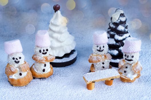 Little snowmen cakes and chocolate trees in powdered sugar sprinkled on the surface. Tinted glass background in cool colours, bokeh.