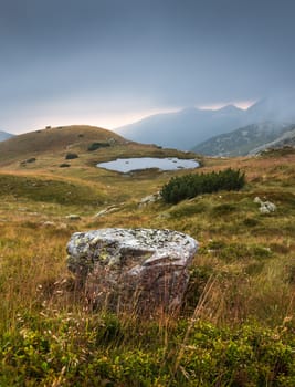 Foggy Mountain Landscape with a Tarn and a Rock in Foreground at Sunset