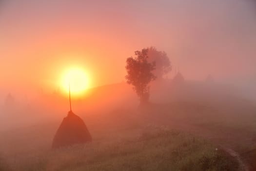 Sunrise over mountain field. Haystacks in misty rural hills. Foggy autumn morning in mountains.