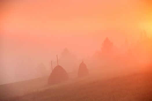 Sunrise over mountain field. Haystacks in misty rural hills. Foggy autumn morning in mountains.