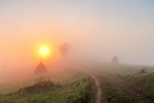 Sunrise over mountain field. Haystacks and road in misty rural hills. Foggy autumn morning in mountains.